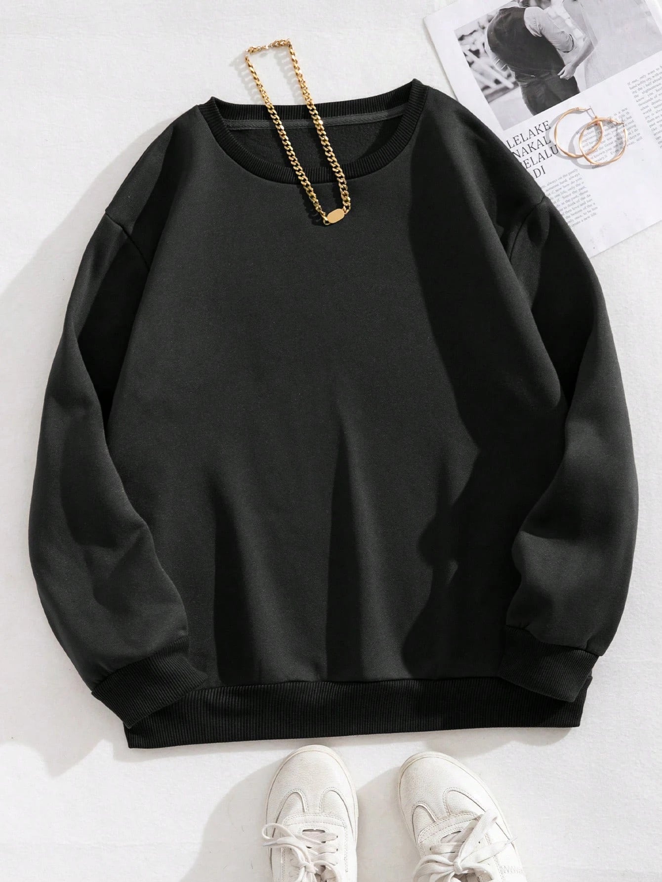Black sweatshirt on a white surface with a gold chain and sneakers.